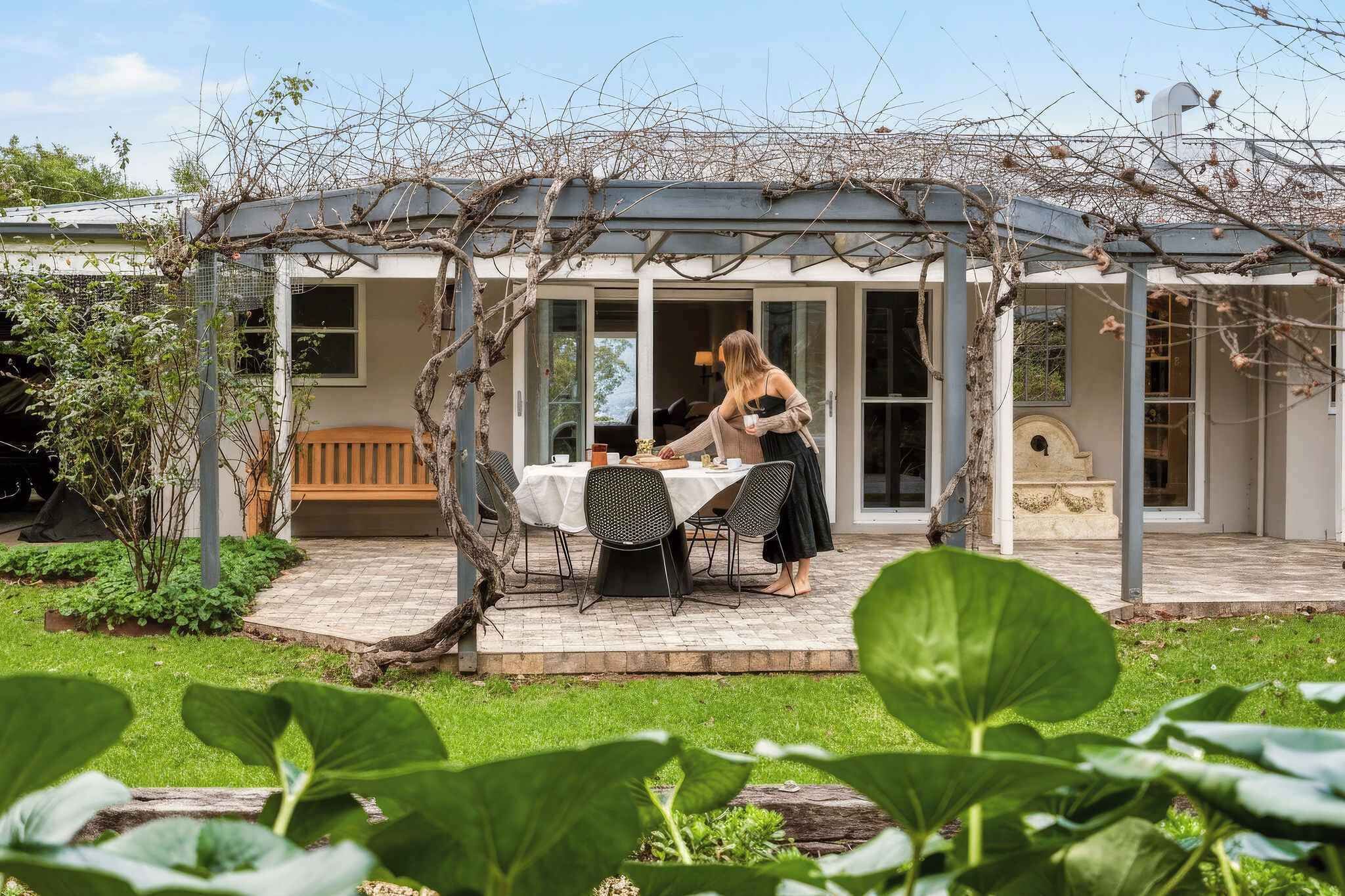 Ein überdachter Essbereich im Freien mit Tisch und Stühlen, umgeben von einem Garten mit verschiedenen Pflanzen.