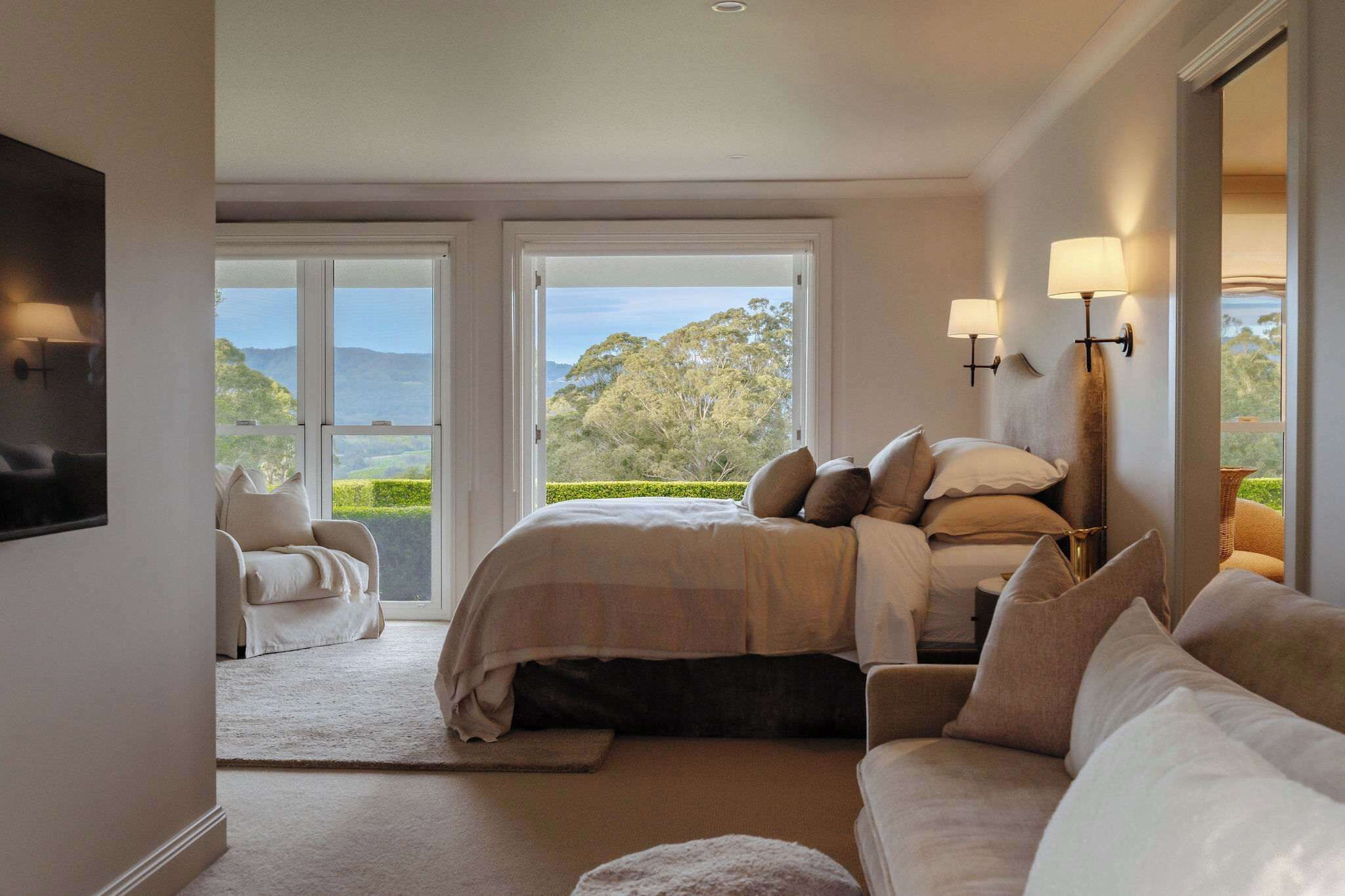 A bedroom with a bed, a sofa, and a television. There are two windows with a view of the outdoors.