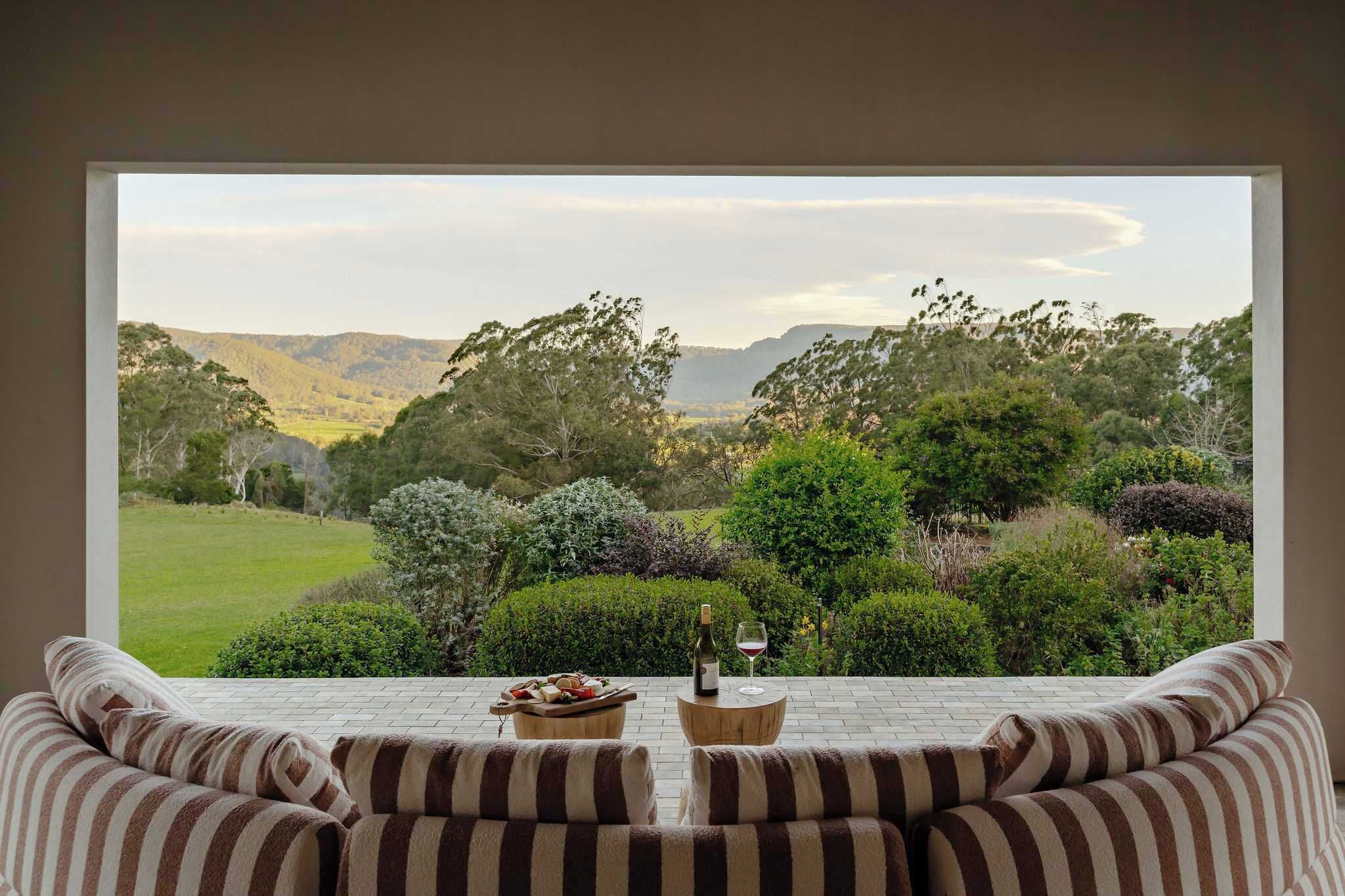 A room with a large window overlooking a landscape with trees and hills. There is a striped sofa and a small table with a bottle and glass.