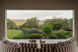 A room with a large window overlooking a landscape with trees and hills. There is a striped sofa and a small table with a bottle and glass. - 'Wychwood'- English Garden Estate with Pool (Upper Kangaroo River)