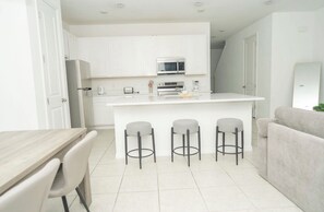 Cocina moderna con armarios blancos, una isla central con taburetes y un sofá gris en una habitación luminosa.