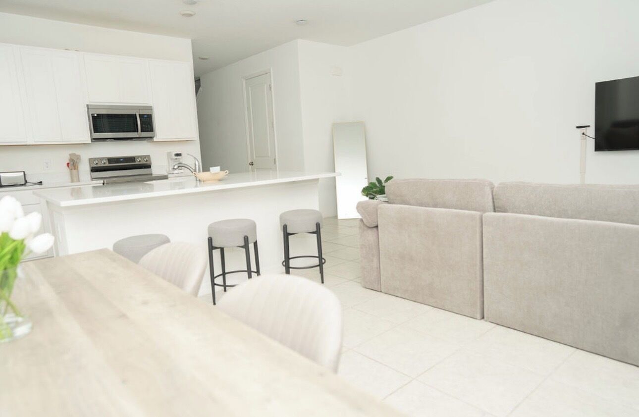 A modern living room with a sofa, a dining table with chairs, and a kitchen area with white cabinetry and appliances.
