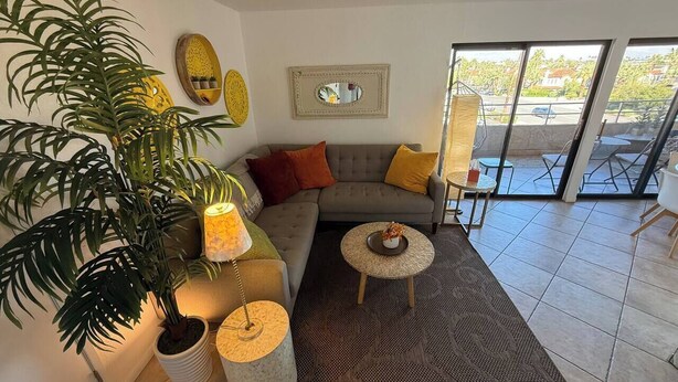 A living room with a sofa, a coffee table, a large potted plant, and a view through a glass door. - Big Beautiful Bed, View and Balcony! (Palm Springs)