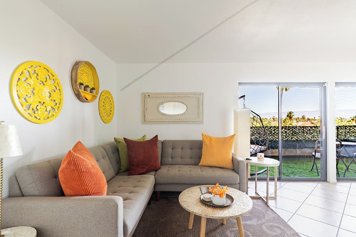Un salon avec un canapé gris, des coussins orange et bruns, une table basse ronde ornée d’un vase, et une grande fenêtre donnant sur un paysage verdoyant.