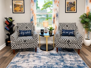 Une pièce comprenant deux fauteuils à motifs, une petite table ronde avec une lampe, un tapis bleu et une bibliothèque.