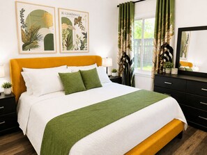 Un lit recouvert d’une couvre-lit verte et blanche, avec une tête de lit de couleur moutarde et deux oreillers verts. Deux reproductions encadrées de motifs botaniques sont accrochées au mur au-dessus du lit.