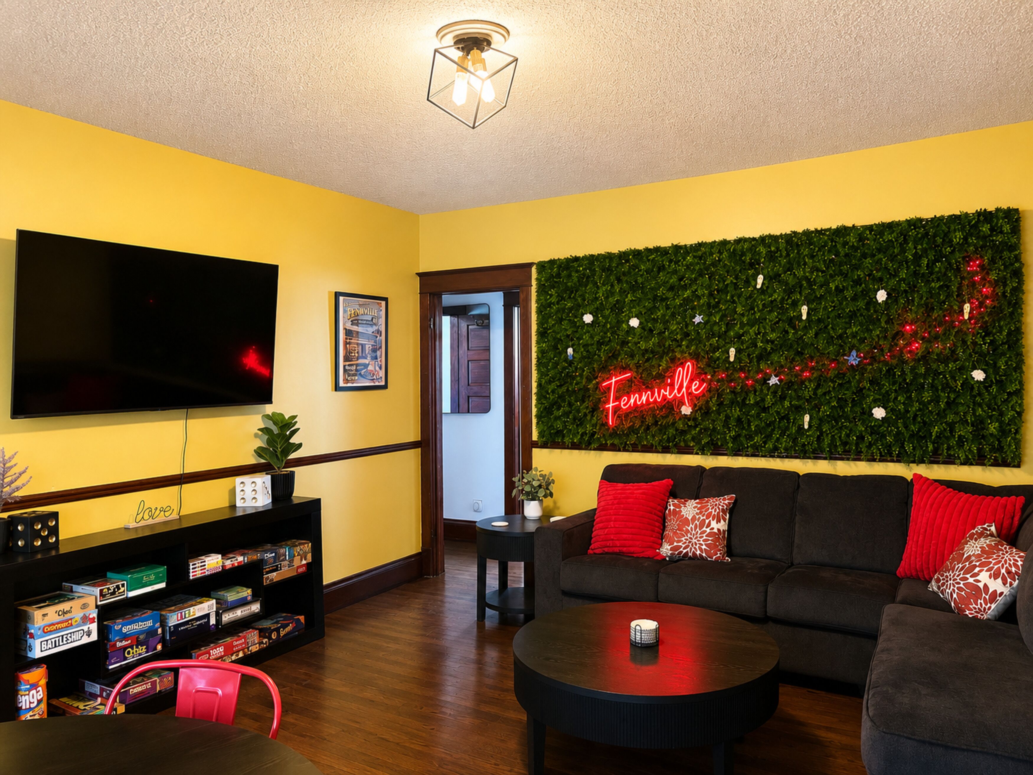 A living room with a flat-screen TV, a green wall with a neon sign, a sofa with red cushions, and a coffee table.