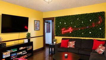 A living room with a flat-screen TV, a green wall with a neon sign, a sofa with red cushions, and a coffee table.
