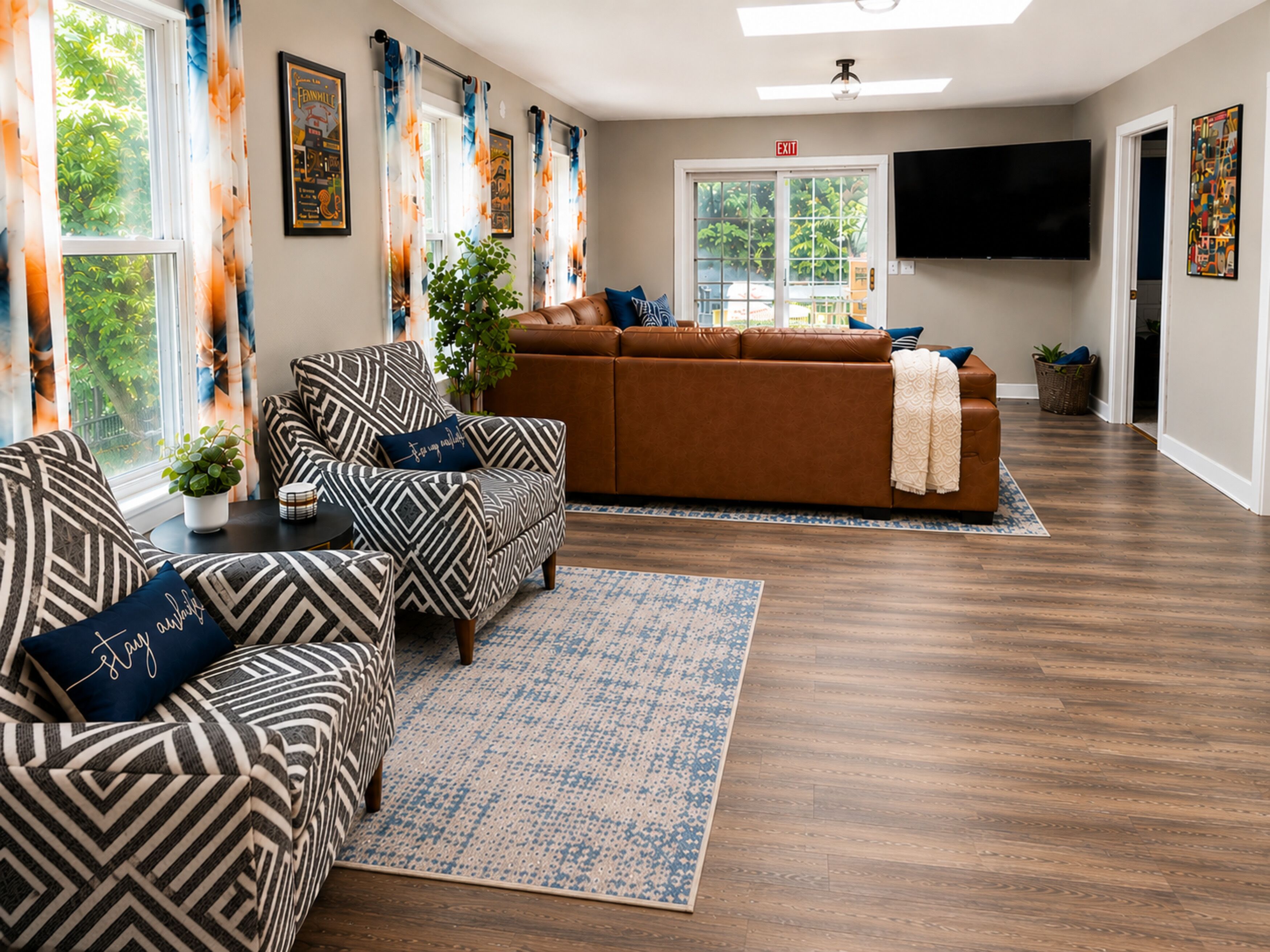 Ein Wohnzimmer mit braunem Sofa, gemusterten Sesseln, einem Flachbildfernseher und einem großen Fenster mit Vorhängen.
