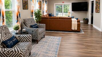 A living room with a brown sofa, patterned armchairs, a flat-screen TV, and a large window with curtains.