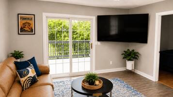 Una sala de estar con un sofá, un televisor de pantalla plana, una mesa de centro redonda y un ventanal con vistas a la vegetación.