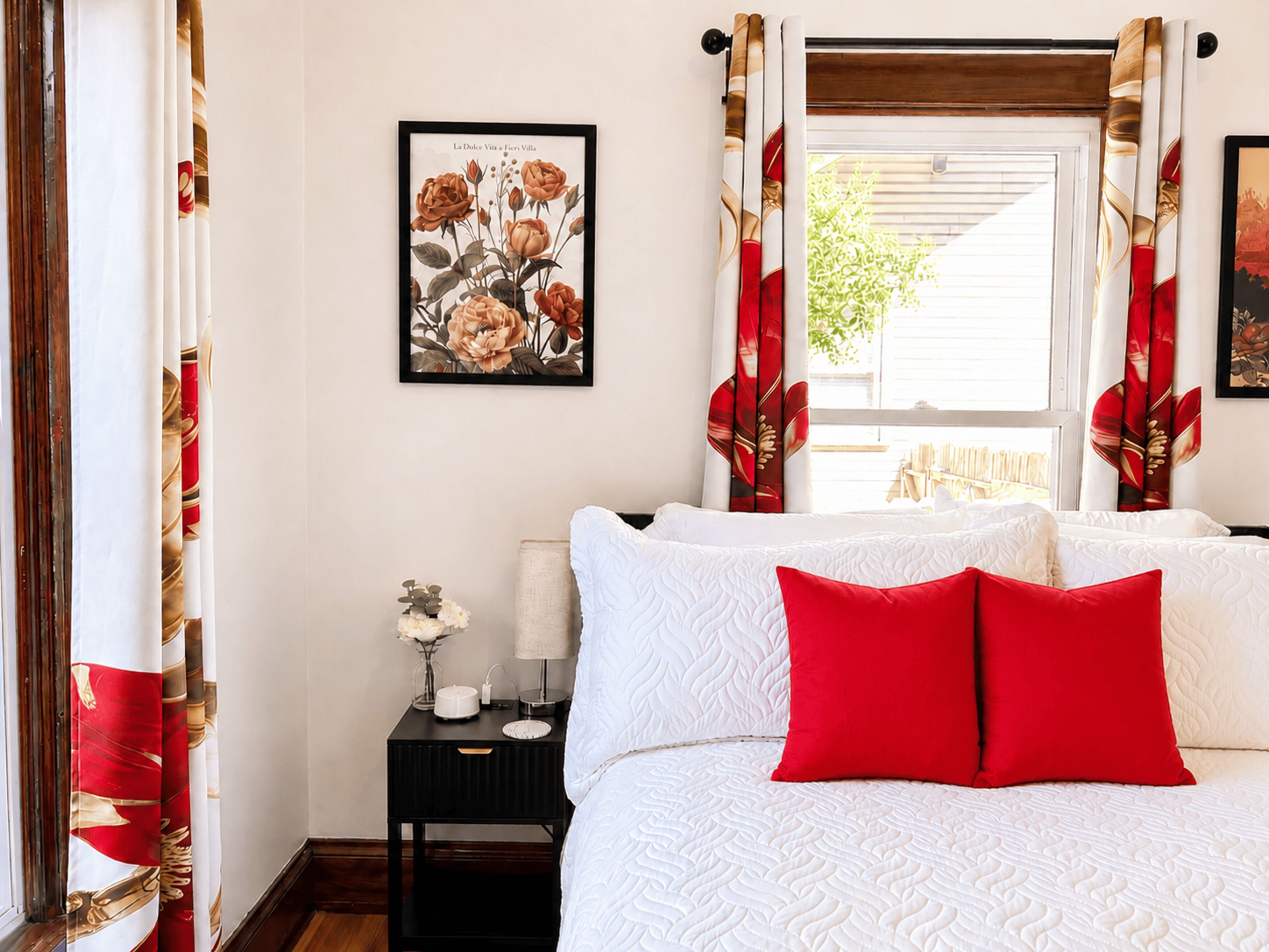 Ein Schlafzimmer mit einem Bett, Kissen, einem Nachttisch, einer gerahmten Blumenillustration und Vorhängen.
