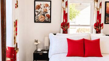 Un dormitorio con cama, almohadas, mesita de noche, una obra de arte floral enmarco y cortinas.