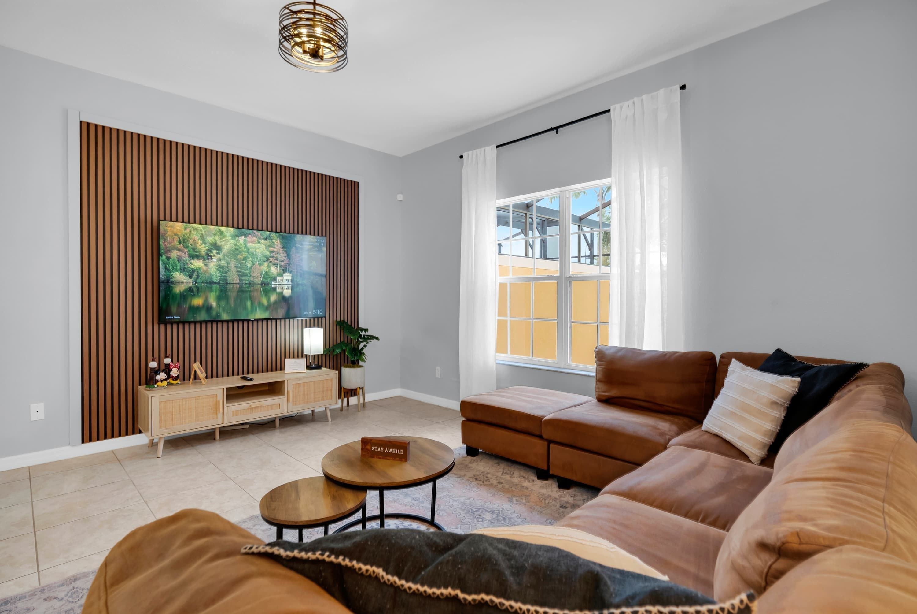 Un salon moderne avec un grand téléviseur à écran plat fixé sur un mur recouvert de panneaux en bois, un canapé en cuir marron, une petite table basse et une fenêtre avec des rideaux.