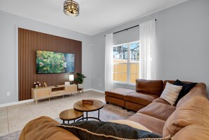 A modern living room with a large flat-screen TV mounted on a wooden panel wall, a brown leather sofa, a small coffee table, and a window with curtains. - Paradise Palms Family Oasis (Kissimmee)