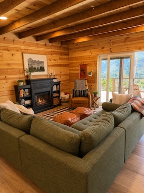A cozy log cabin interior with a sectional sofa, a fireplace, and a view of the outdoors.