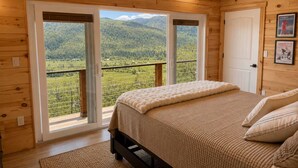 Een slaapkamer in een houten chalet met een bed, een plafondventilator en uitzicht op een bebost berglandschap door grote ramen.