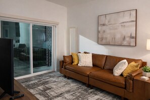A living room with a brown leather sofa, a flat-screen TV, a remote control, and a patterned rug. - Modern 1BR Near Downtown Atlanta Pool, Gym & Balcony (Atlanta)