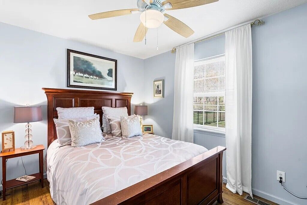 A bedroom with a wooden bed, a ceiling fan, a window with curtains, and a framed picture on the wall.