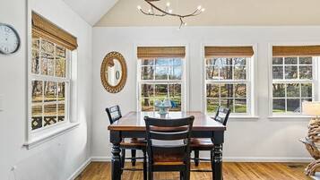 A dining room with a wooden table and chairs, large windows with blinds, a wall clock, and a chandelier.
