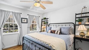 Un dormitorio con cama, ventilador de techo, ventana con cortinas, mesita de noche con lámpara y estante con objetos decorativos.