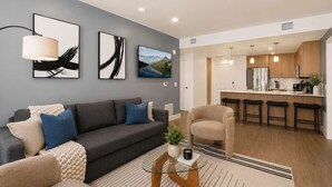 Uma sala de estar moderna com um sofá cinzento, uma mesa de centro e uma poltrona. Há uma área de cozinha com armários de madeira e banquetas.