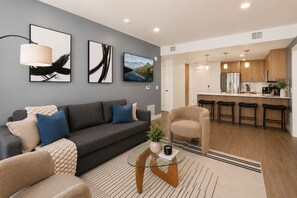 Ein modernes Wohnzimmer mit grauem Sofa, einem Couchtisch und einem Sessel. Es gibt eine Küchenzeile mit Holzschränken und Barhocker.