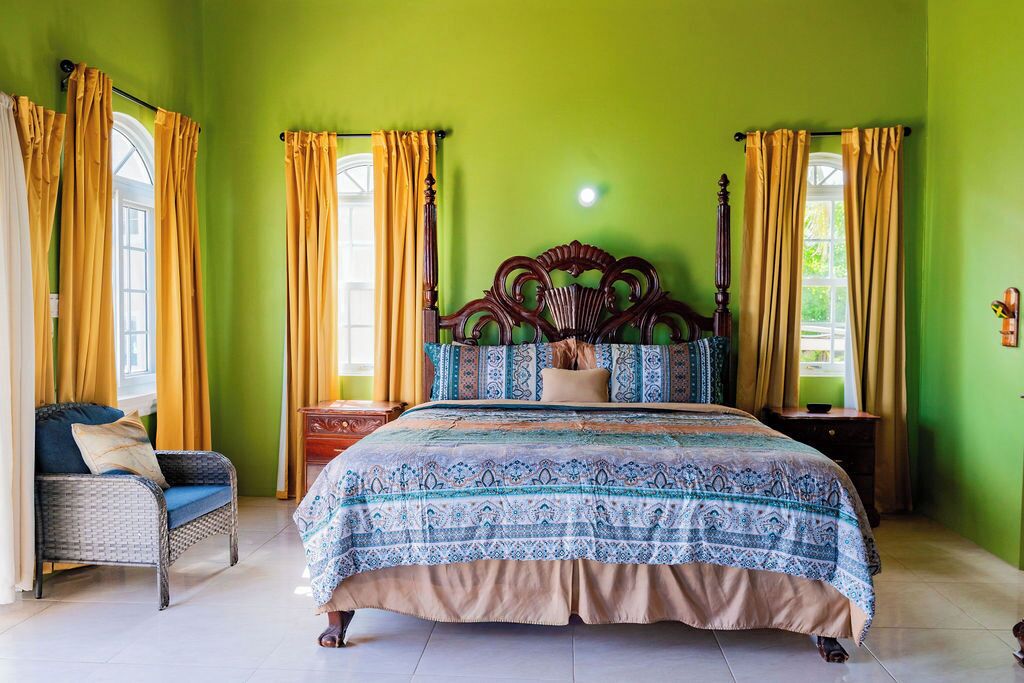 A bedroom with a large bed, a wooden headboard, a wicker chair, and curtains on the windows.