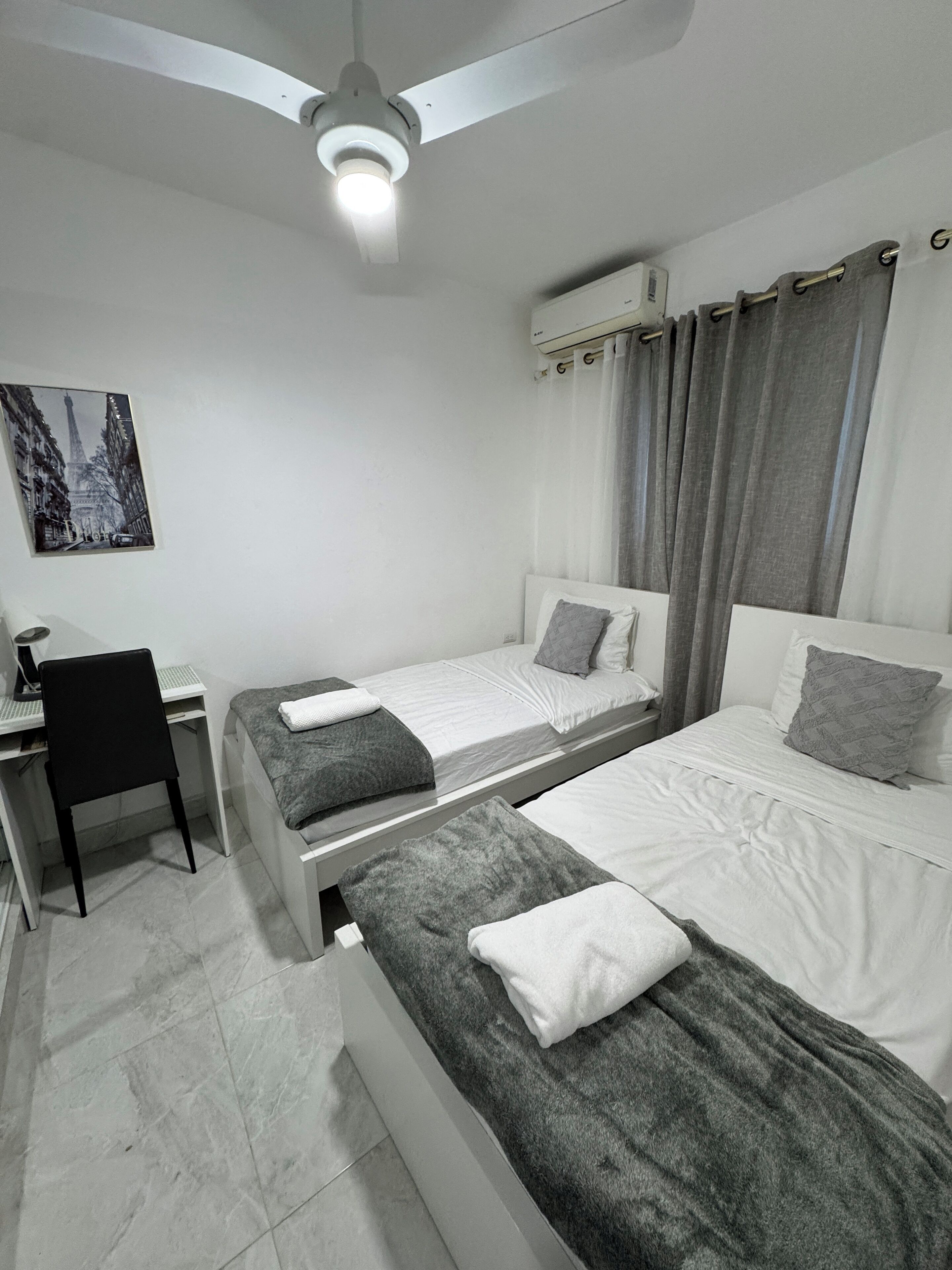 Ein Schlafzimmer mit zwei Betten, einem Schreibtisch, einem Stuhl, einem Deckenventilator und einer Klimaanlage.