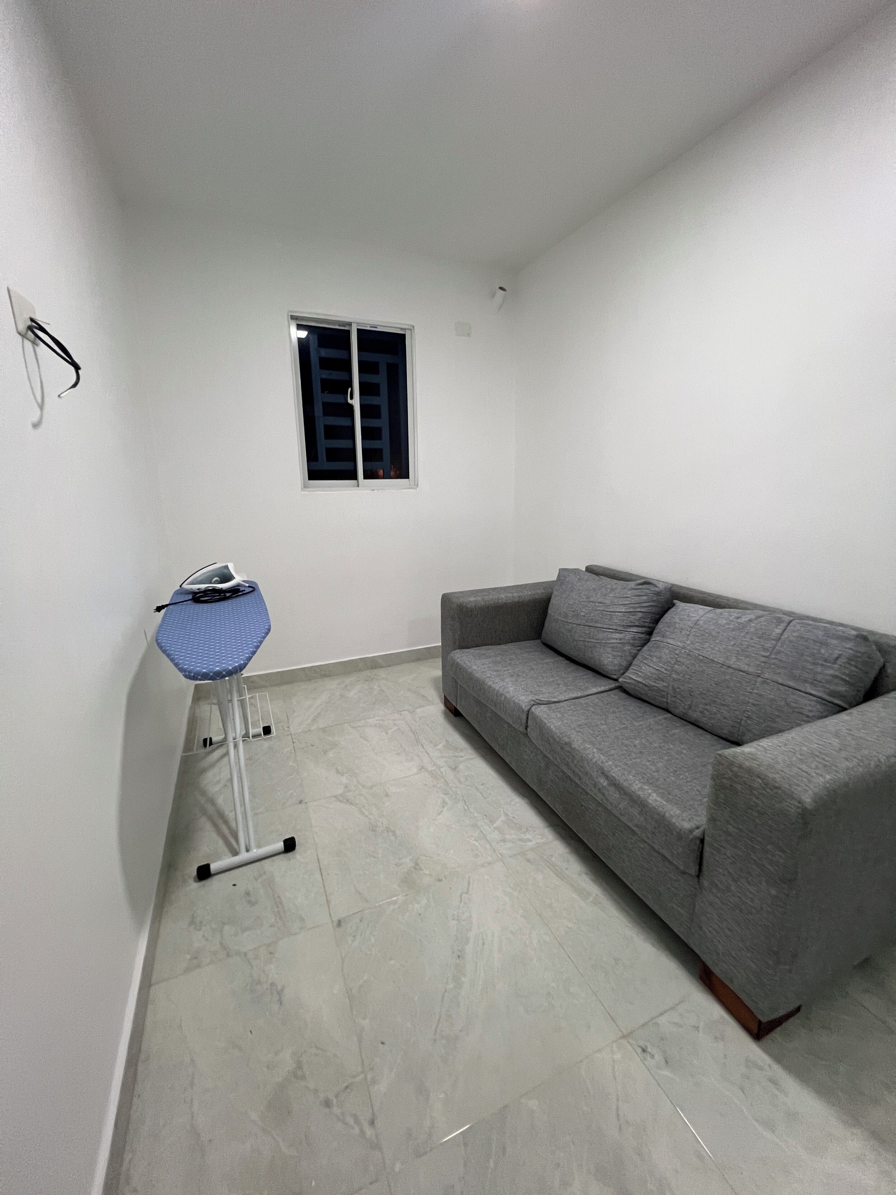 Una habitación con un sofá gris, una silla plegable azul, un ventanal pequeño y una lámpara de pared.
