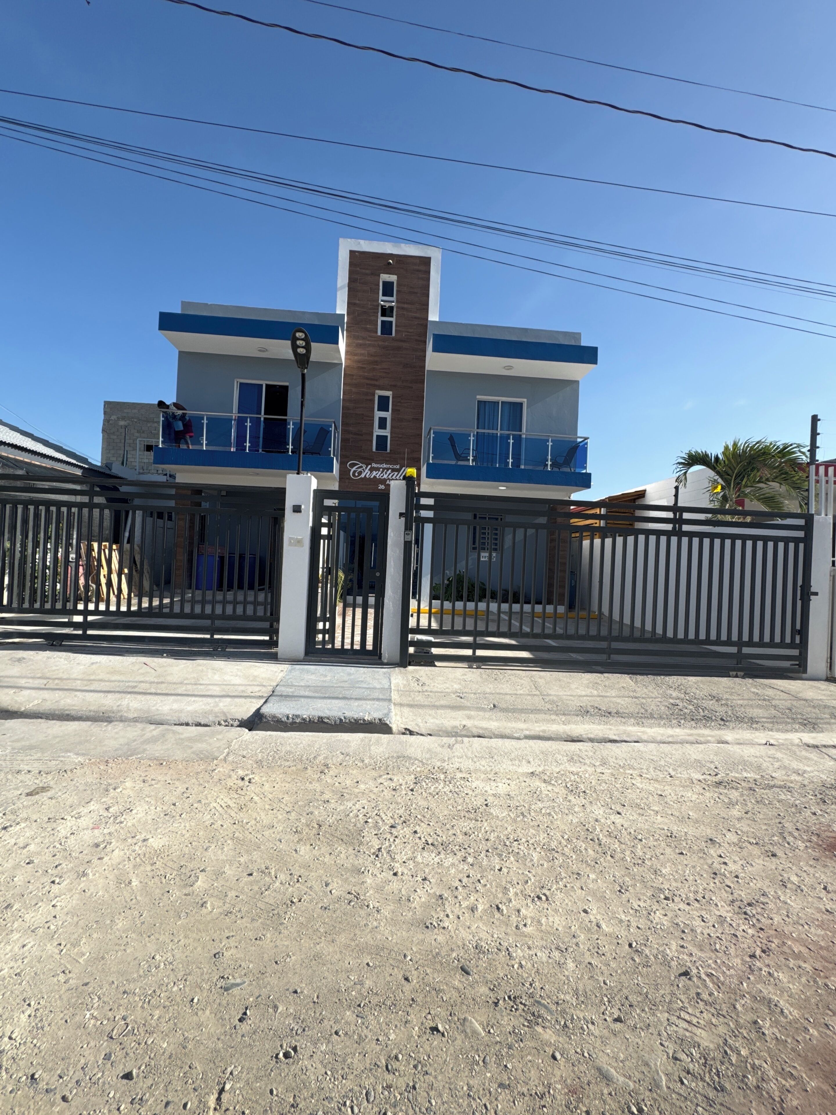 A two-story building with a gated entrance, a sign indicating 'Residencial Cristal 20', and a clear blue sky.