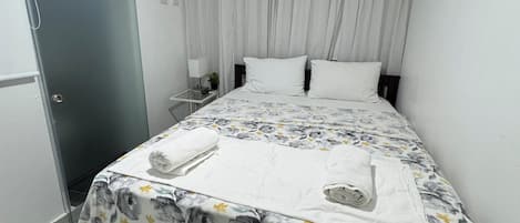 A bedroom with a bed, curtains, an air conditioning unit, a bedside table, and a wall-mounted picture.