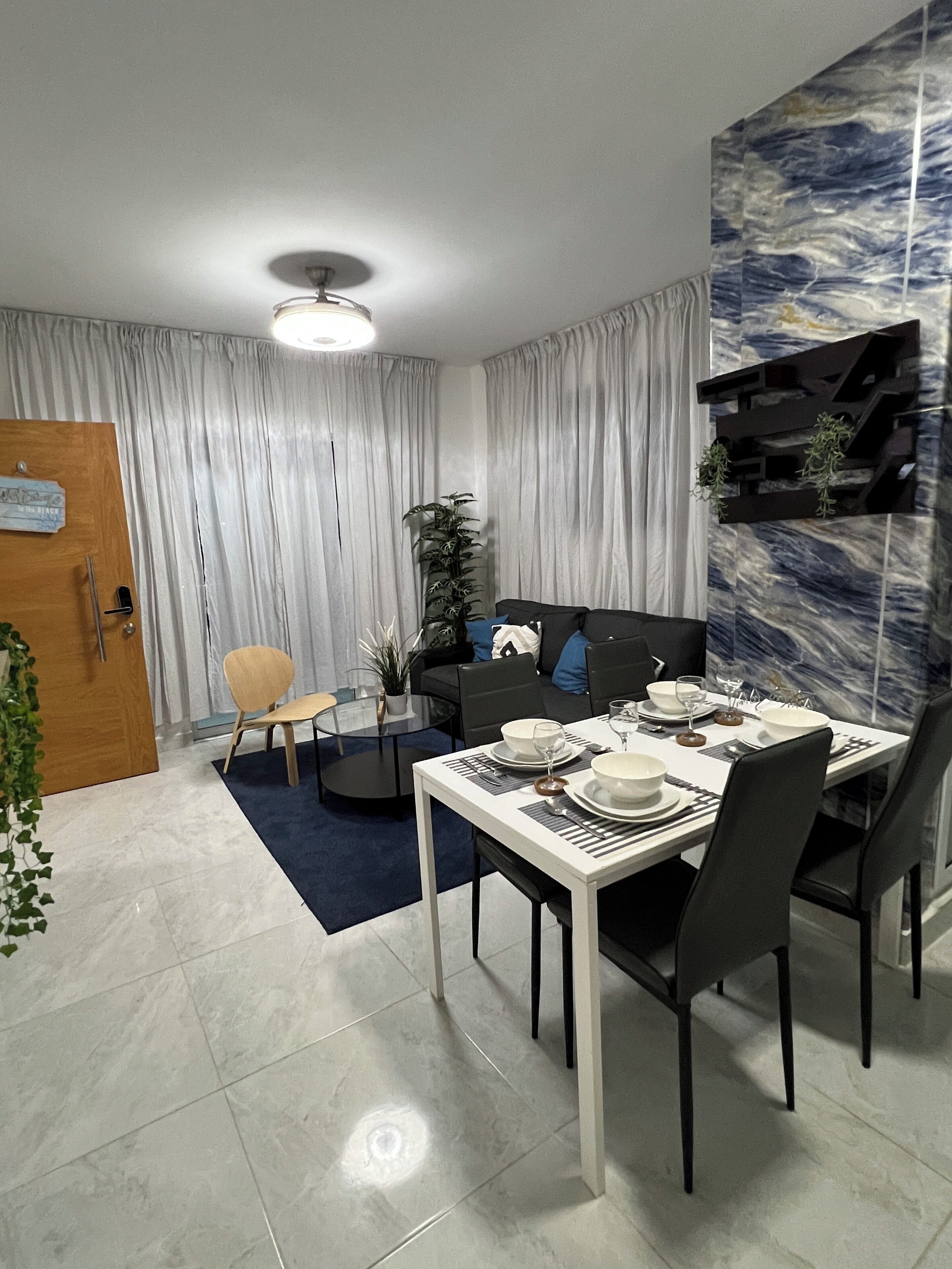 Un comedor moderno con una mesa blanca dispuesta para cuatro comensales, un sofá de color oscuro y una mesa de centro de cristal.