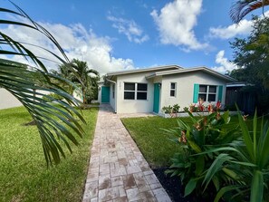 A modern two-story house with a paved driveway leading to the front door, surrounded by well-maintained grass and tropical plants. - Sunny 2BR Apartment w/ Private Backyard & Hot Tub (Wilton Manors)