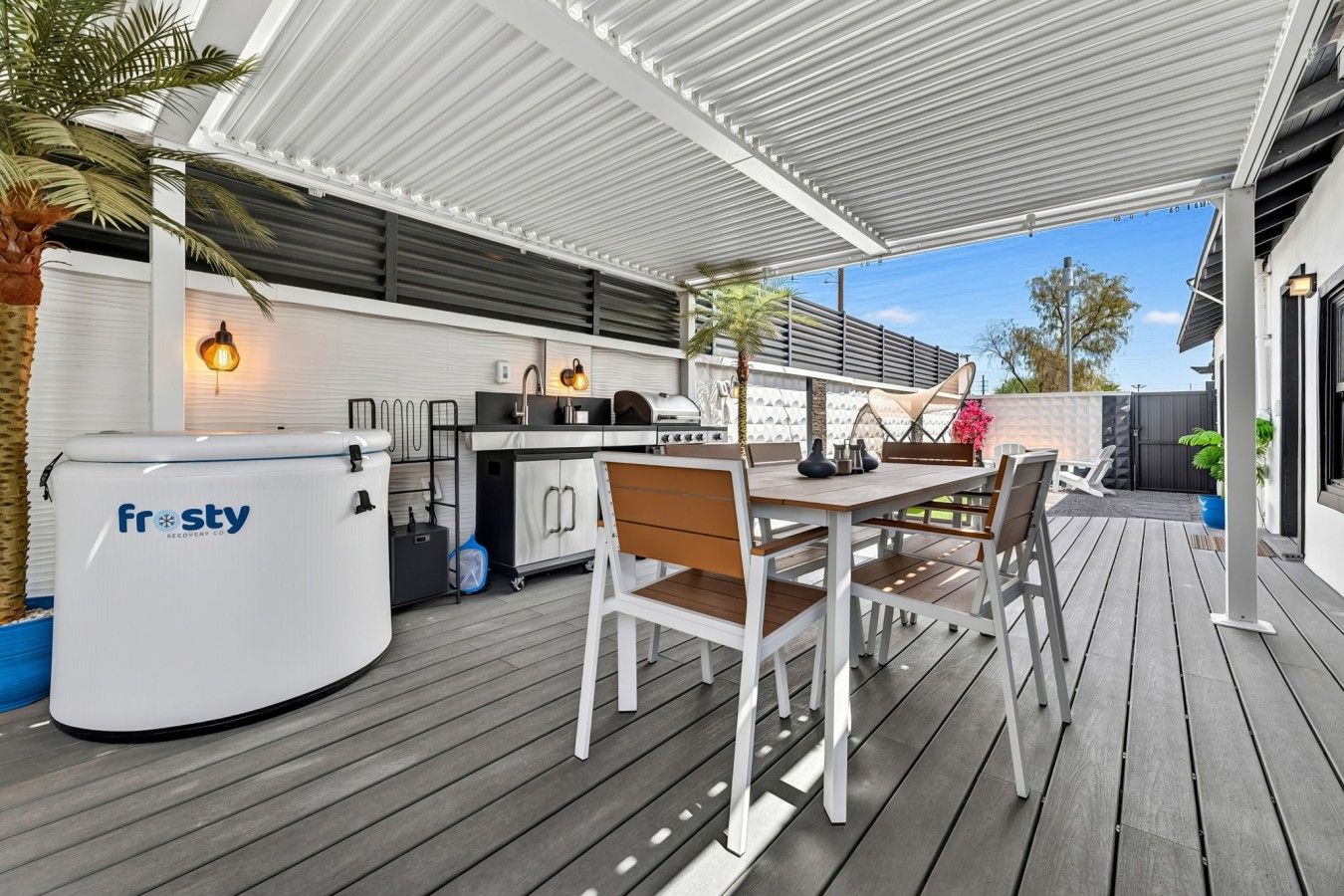 Une terrasse extérieure dotée d’une plateforme en bois, d’une cuisine extérieure blanche équipée d’un chauffe-eau de la marque Frosty, et d’un espace détente avec des chaises et une table.