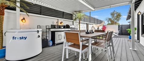Une terrasse extérieure dotée d’une plateforme en bois, d’une cuisine extérieure blanche équipée d’un chauffe-eau de la marque Frosty, et d’un espace détente avec des chaises et une table.