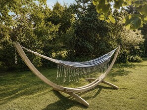 Een hangmat, opgehangen tussen twee houten palen, in een grasveld met weelderige begroeiing op de achtergrond.