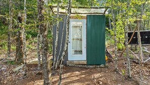 Eine kleine, rustikale Holzhütte mit Tür und Fenster, umgeben von Bäumen und einer Holzterrasse.