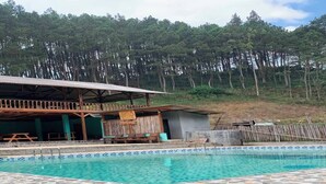 A swimming pool with a tiled edge, a wooden deck, and a building with a balcony in the background. - Cozzy Taman Nyingkur (bandung)