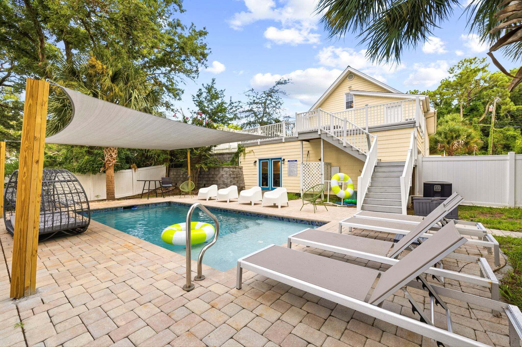 A backyard with a swimming pool, lounge chairs, a covered patio, and a house with a balcony.