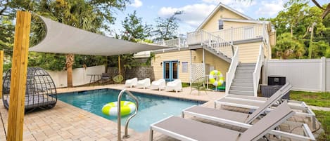 A backyard with a swimming pool, lounge chairs, a covered patio, and a house with a balcony.