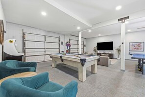 Ein modernes Wohnzimmer mit Billardtisch, smaragdgrünen Sesseln, einem Fernseher und einer Garage mit weißen Türen.