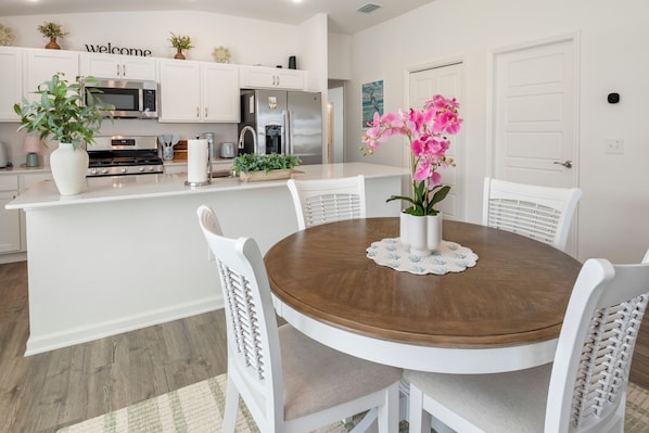 A dining area with a round wooden table, white chairs, and a vase with pink flowers.