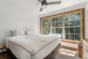 Ein Schlafzimmer mit großem Fenster, einem Bett mit Blumenmuster-Quilt, einem Deckenventilator und einer hölzernen Kommode.