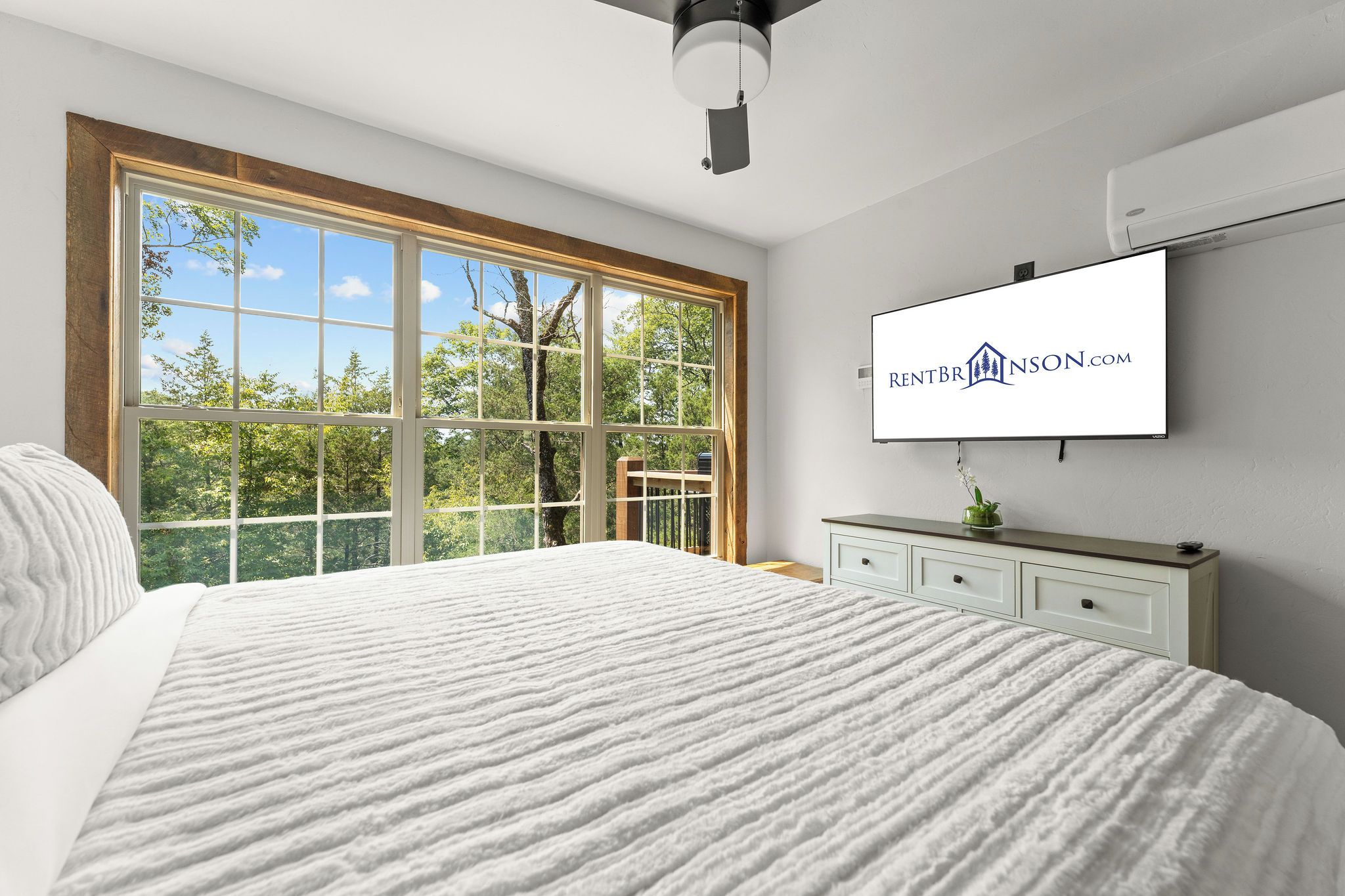 Un dormitorio con un ventanal grande, una cama con un edredón blanco texturizado, un aparador blanco y un televisor de pantalla plana montado en la pared mostrando un logotipo.