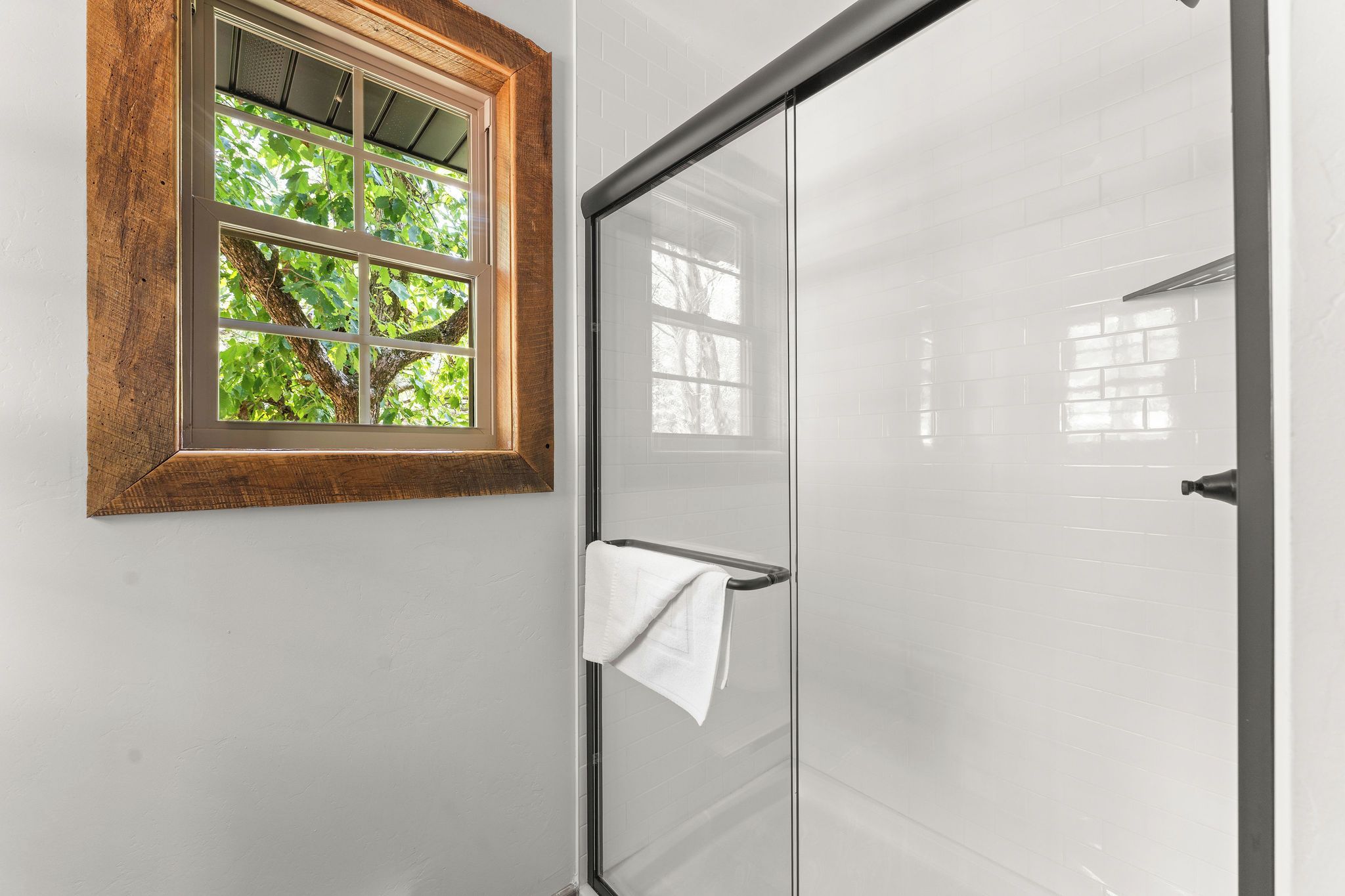 Ein Badezimmer mit einer Duschkabinentür aus Glas, einem Fenster mit Holzrahmen und einem Handtuch, das an der Duschkabinentür hängt.