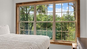 A bedroom with a large window, a bed with a white textured blanket, and a wooden dresser. - Cozy Cabin Tucked Away w Free Attraction Tickets_Deck Space_Bonus Loft_Near Branson (Branson)
