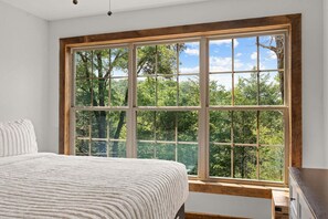 A bedroom with a large window, a bed with a white textured blanket, and a wooden dresser. - Cozy Cabin Tucked Away w Free Attraction Tickets_Deck Space_Bonus Loft_Near Branson (Branson)