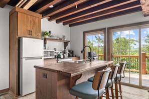 A kitchen with a wooden island, bar stools, a refrigerator, and a view of greenery through large windows. - Tucked Away Cabin w Peaceful Views_Near Branson_Free Attraction Tickets (Branson)