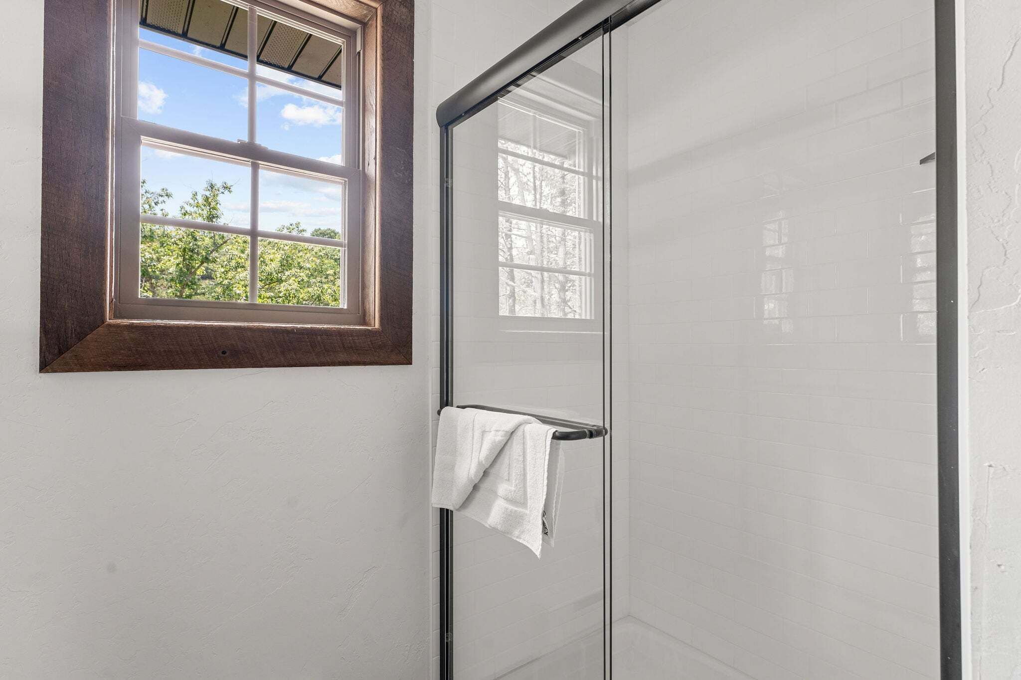 A bathroom with a glass shower door, a towel hanging on a rack, and a window with a wooden frame.